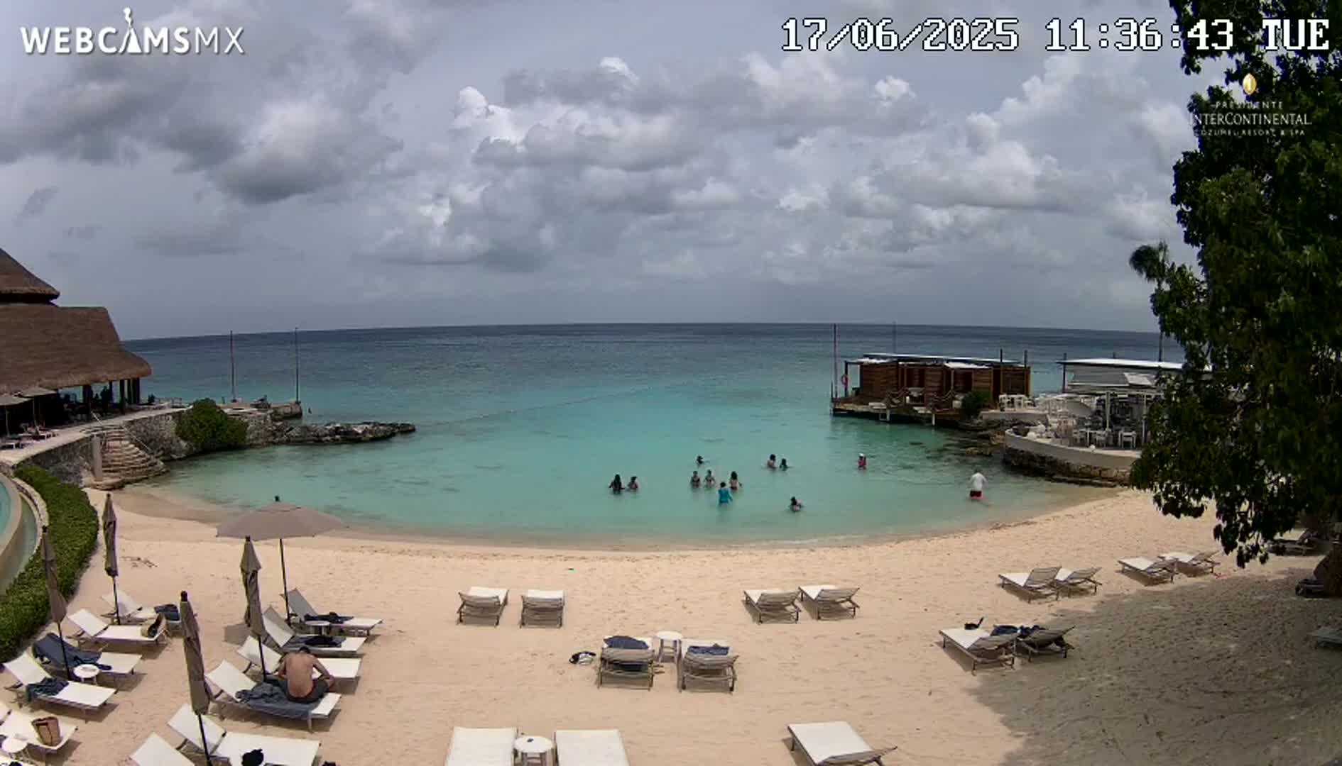A partly cloudy day at a beach with turquoise water, several people swimming, lounge chairs set up on the sand, and a building with a thatched roof.