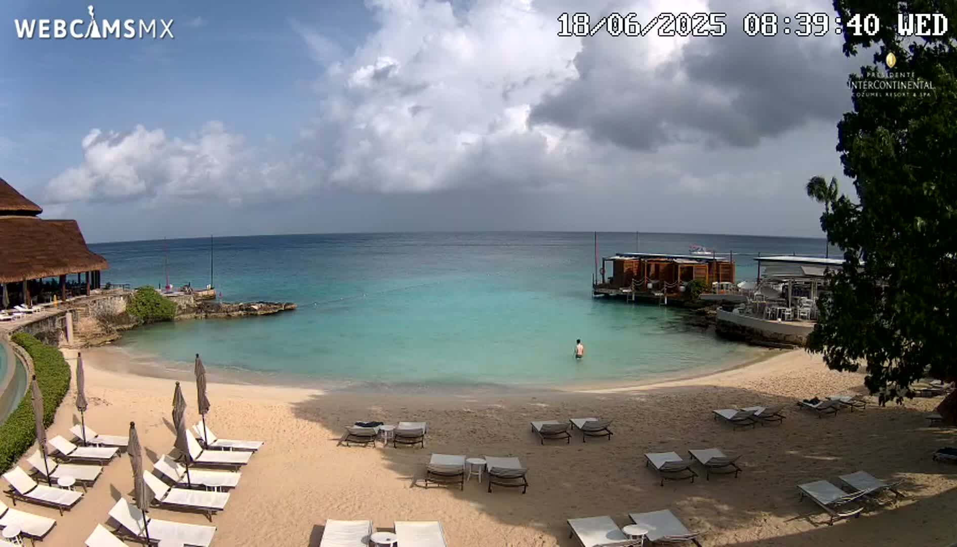 A partly cloudy day at a beach resort shows a sandy beach with lounge chairs, calm turquoise water, and a small overwater structure.