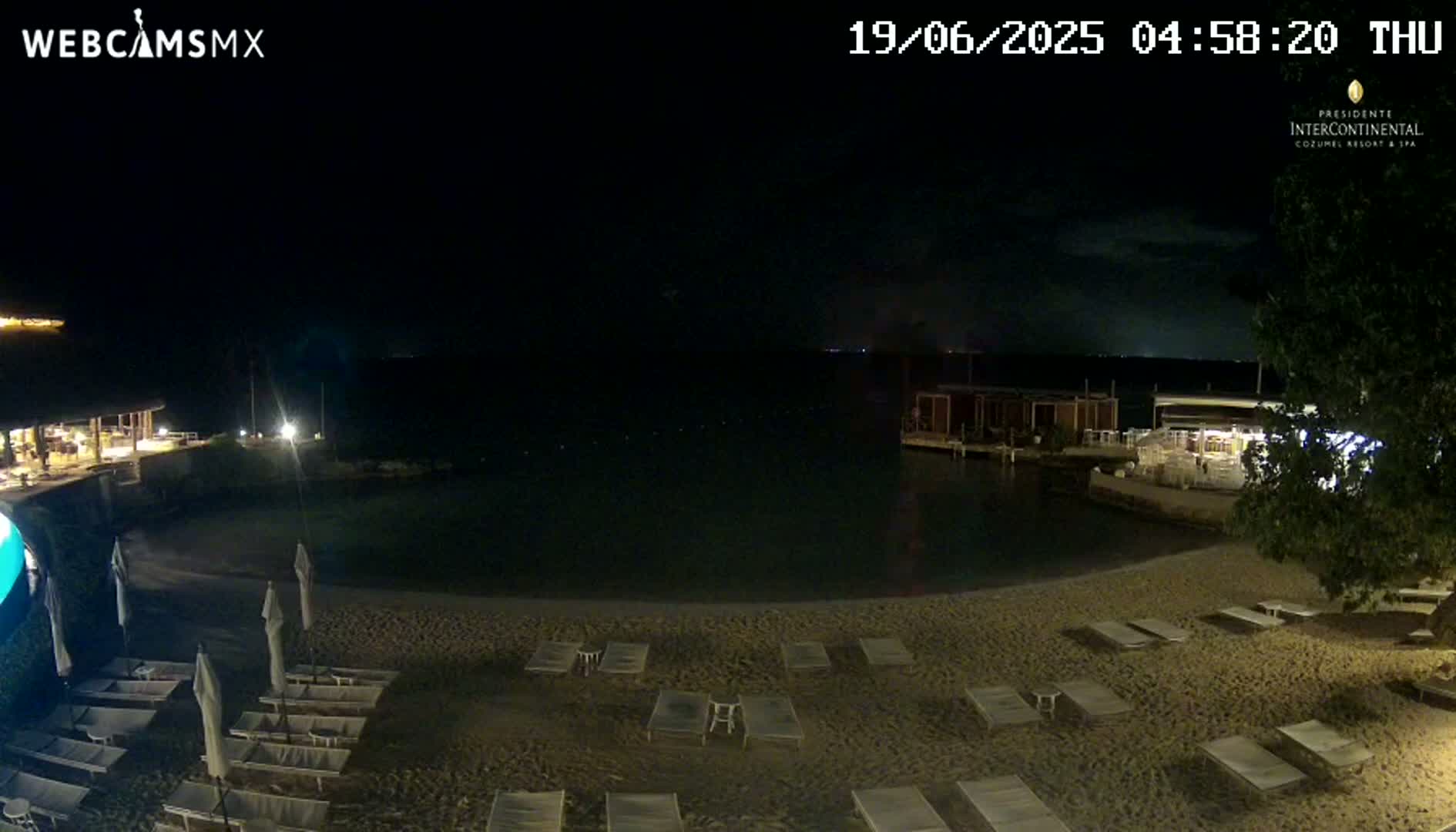 A dark nighttime beach scene shows a sandy shore with lounge chairs, a calm body of water, and buildings lit along the shoreline under a mostly clear sky.