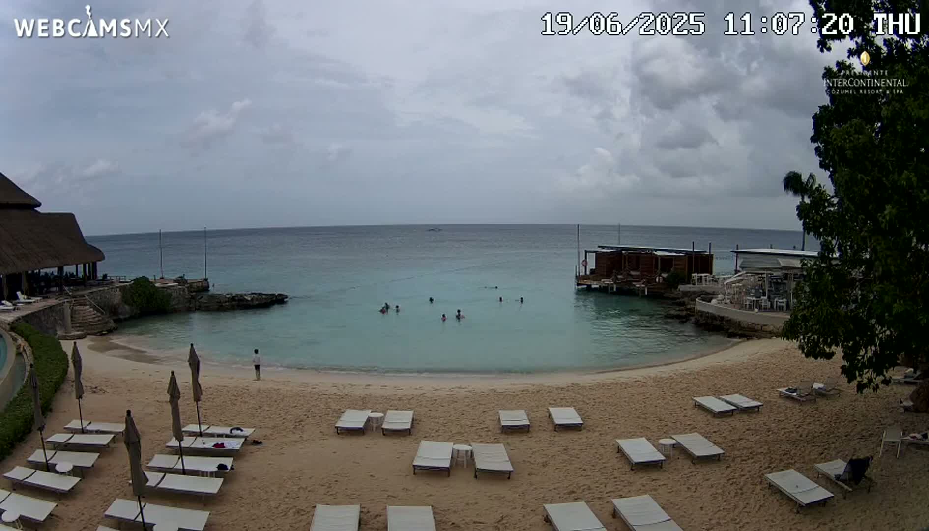 A sandy beach with lounge chairs and people swimming in calm, turquoise water under a partly cloudy sky.