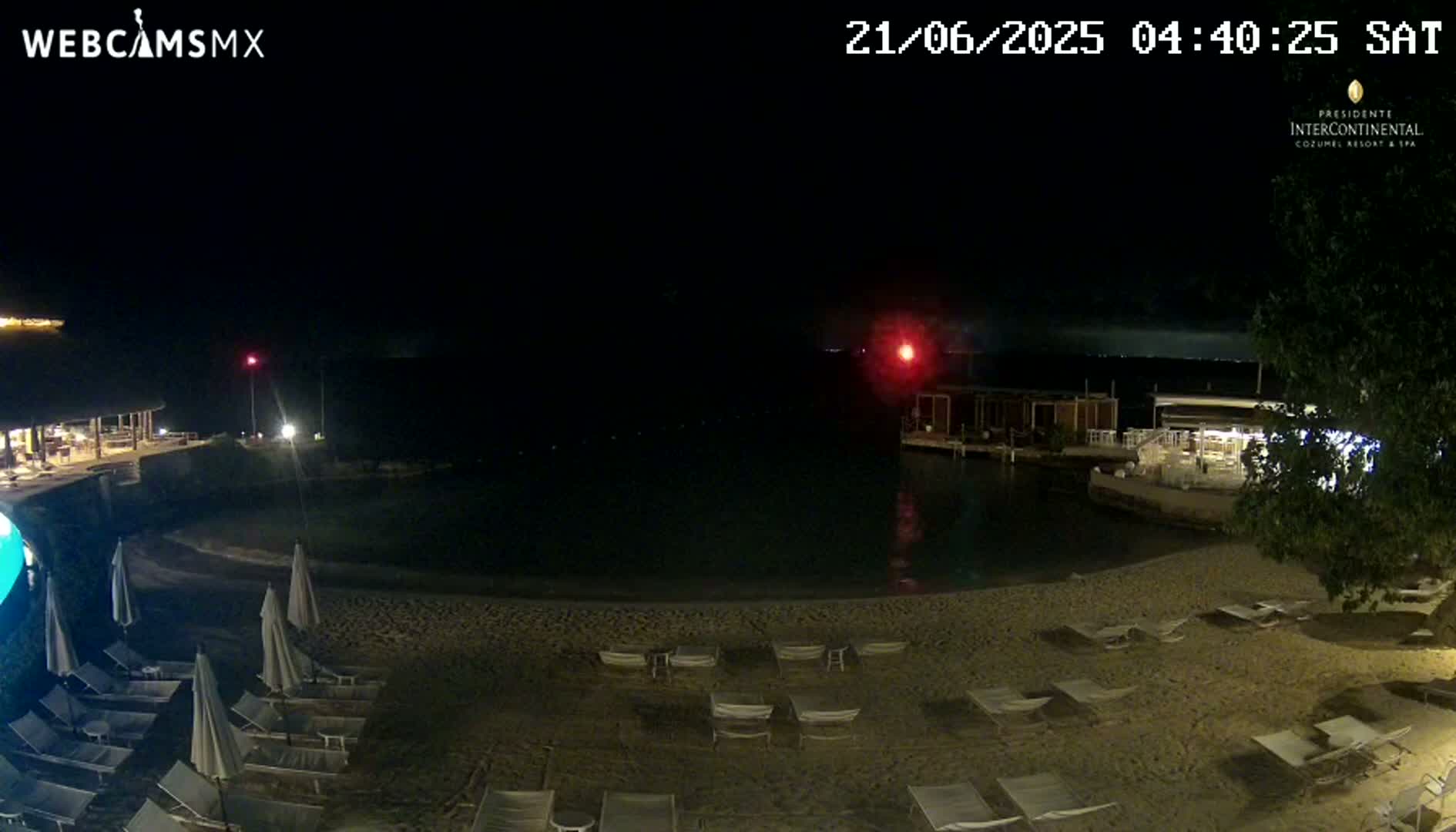 A nighttime beach scene shows empty lounge chairs on a sandy beach next to calm water, with a restaurant visible in the background under a clear, dark sky.