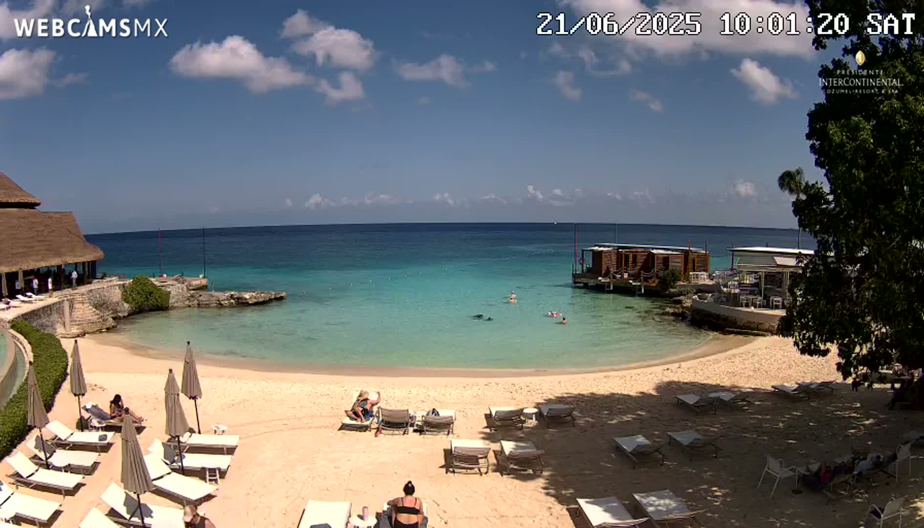 A sandy beach with turquoise water, lounge chairs, and several people relaxing, under a partly sunny sky.