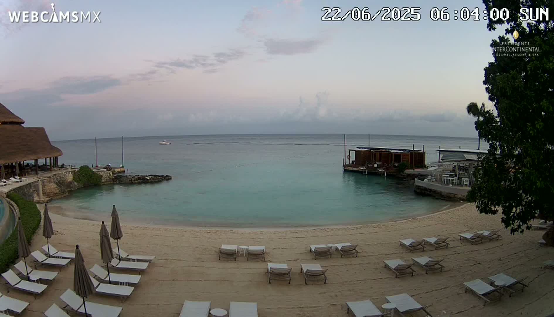 A tranquil beach scene with calm, light-blue water under a mostly cloudy sky shows empty lounge chairs on a sandy shore near a resort building and an overwater structure.