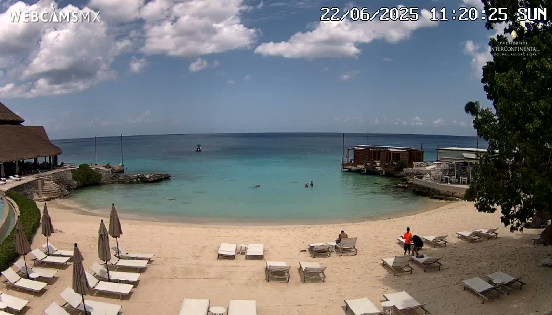 A partly sunny beach scene shows calm, turquoise water, a sandy shore with lounge chairs and umbrellas, and a few people swimming and relaxing.