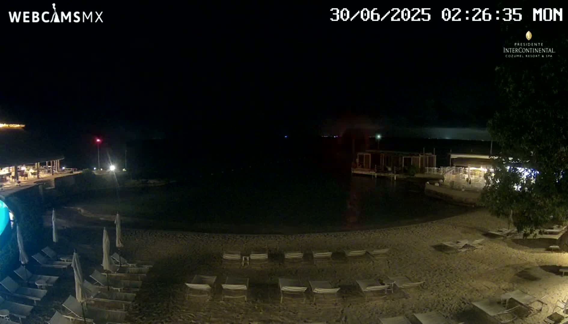 A nighttime view of a sandy beach with lounge chairs, a calm body of water, and some buildings nearby under a dark sky.