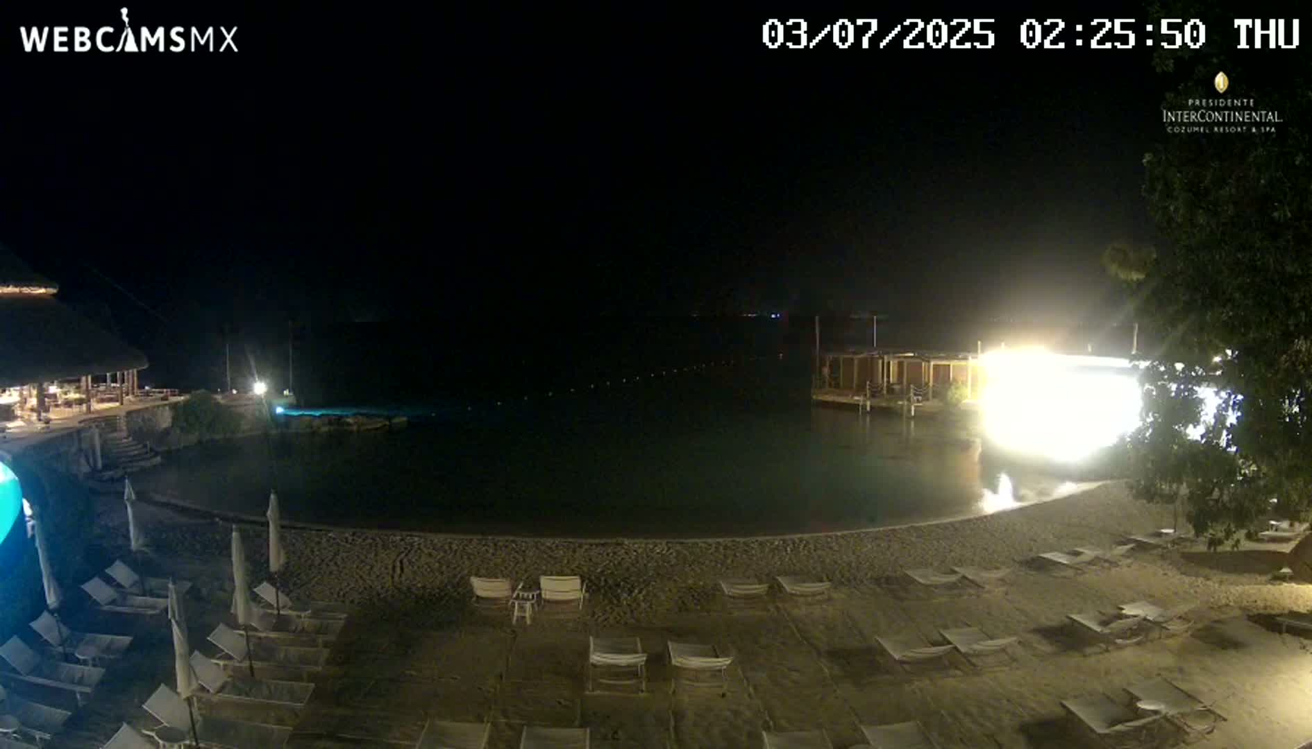 A nighttime view of a calm body of water with a sandy beach lined with lounge chairs, and a building with lights on in the background.