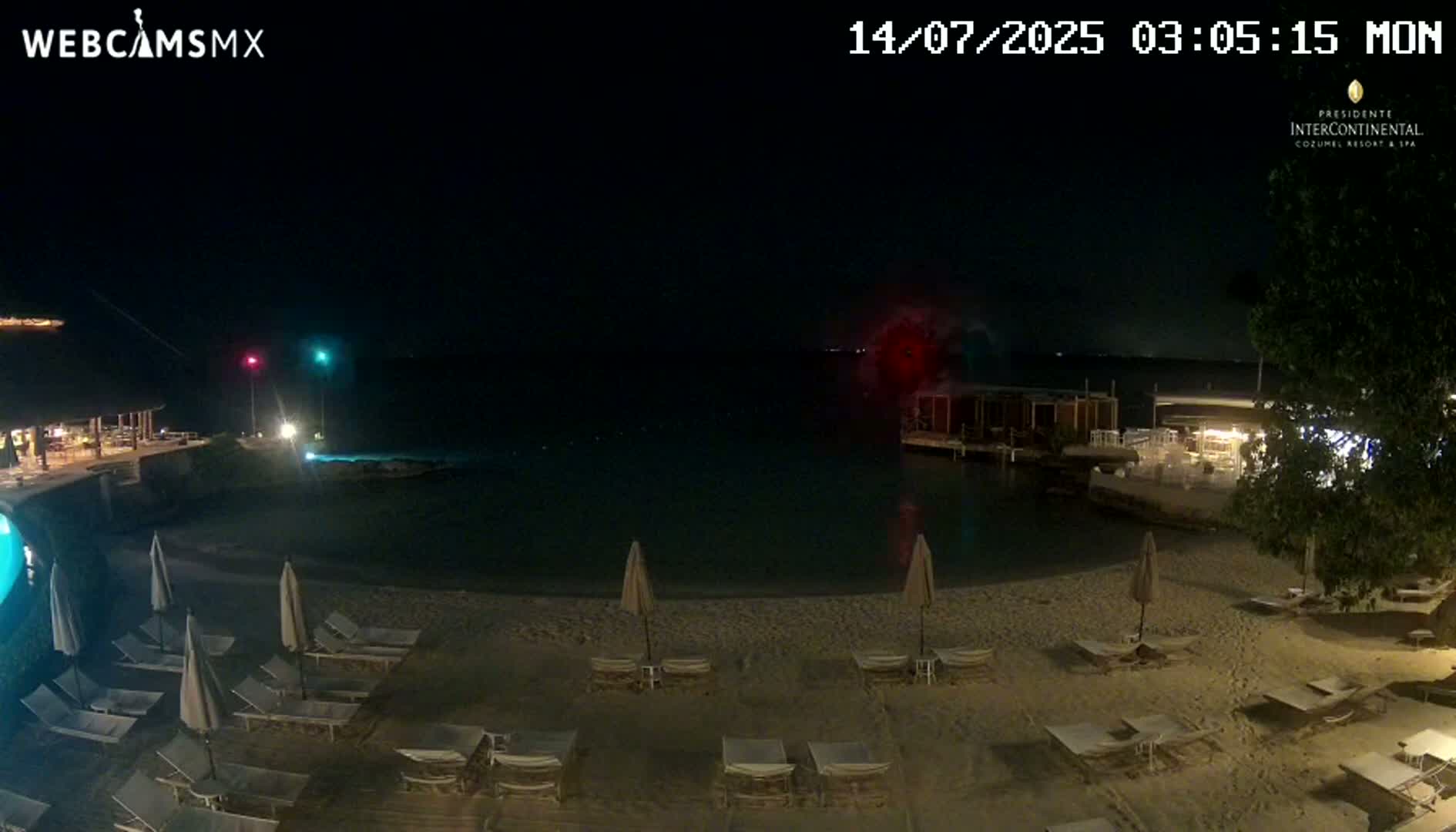 A nighttime beach scene shows calm water, lounge chairs under umbrellas on the sand, and a building with lights across the water.