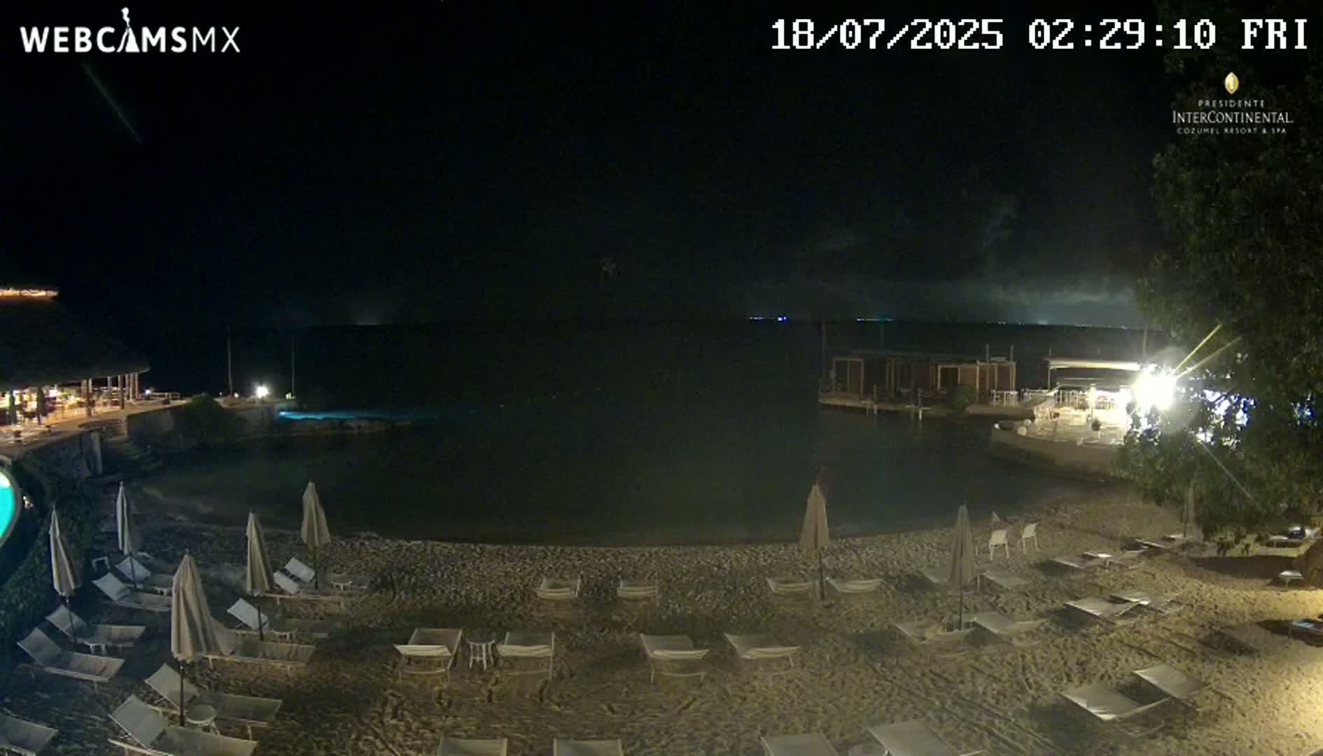 A dark nighttime beach scene shows empty lounge chairs and umbrellas on a sandy beach next to calm water under a clear, dark sky.