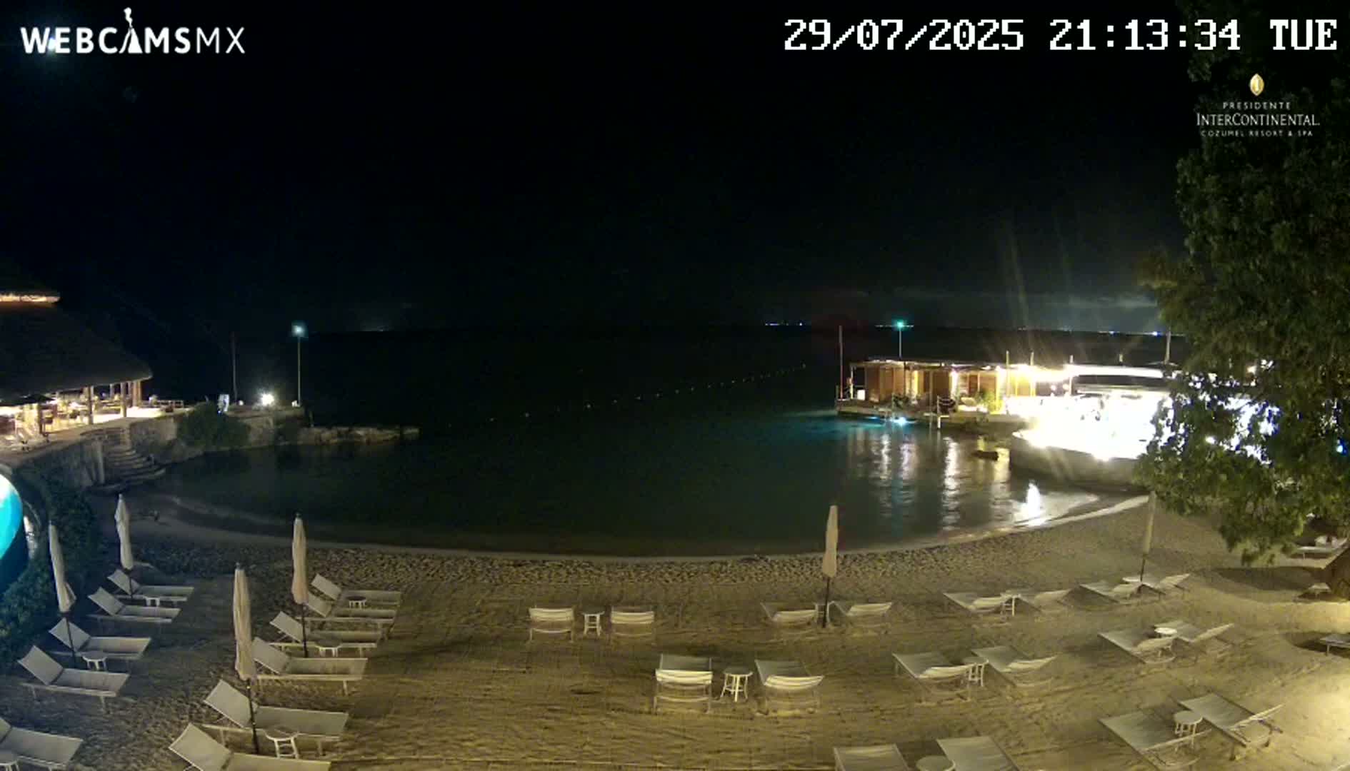A dark nighttime beach scene shows empty lounge chairs and umbrellas on a sandy beach next to calm water under a clear, dark sky.