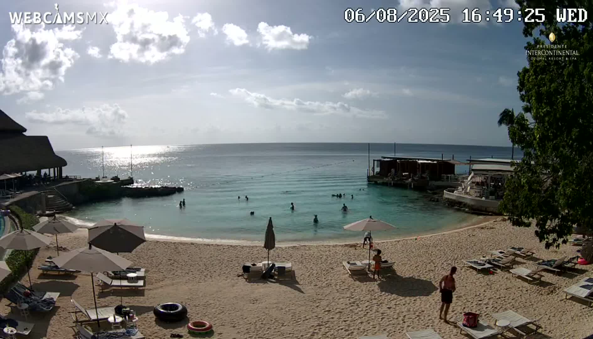 A sandy beach with lounge chairs and umbrellas, calm turquoise water with people swimming, and a partly sunny sky.