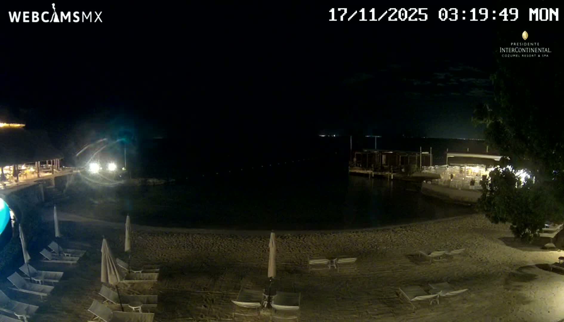 The image captures a resort beach at night, featuring lounge chairs and closed umbrellas on the sand, a lit pier and structures over tranquil dark water, and distant lights under a clear sky.