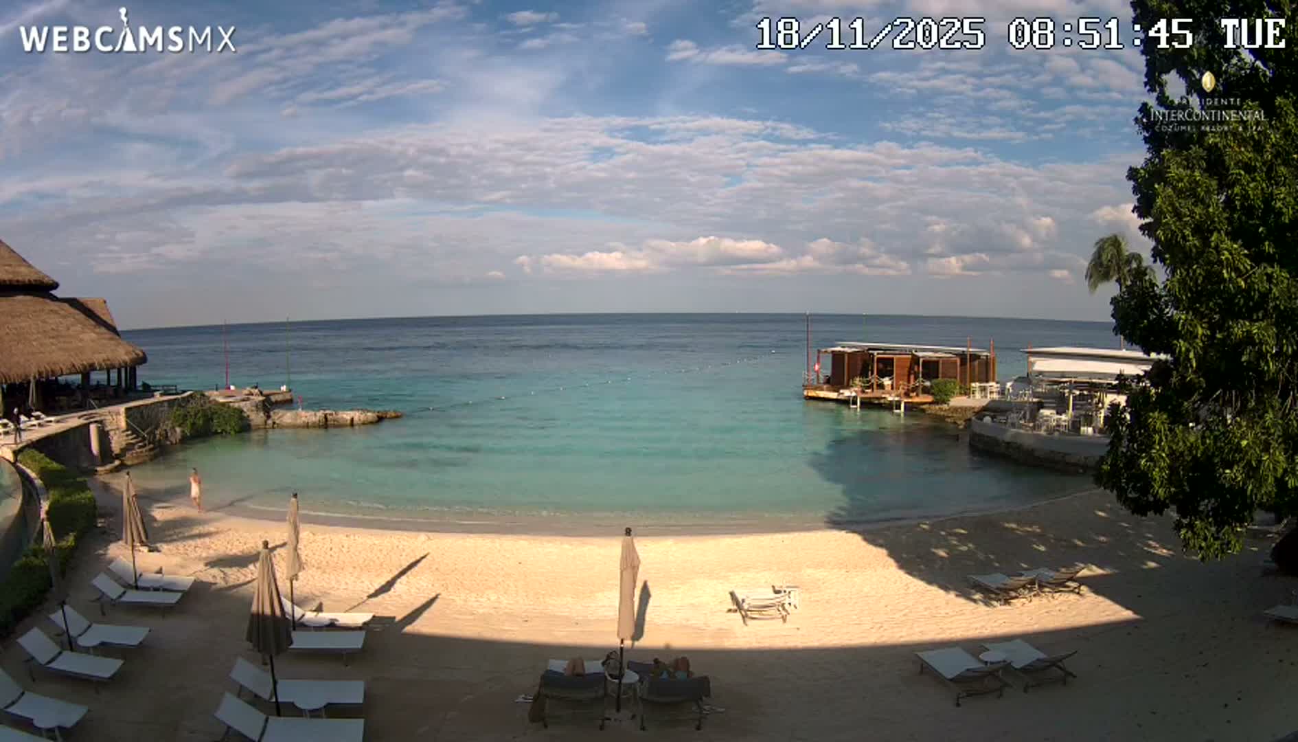 The image captures a resort beach at night, featuring lounge chairs and closed umbrellas on the sand, a lit pier and structures over tranquil dark water, and distant lights under a clear sky.