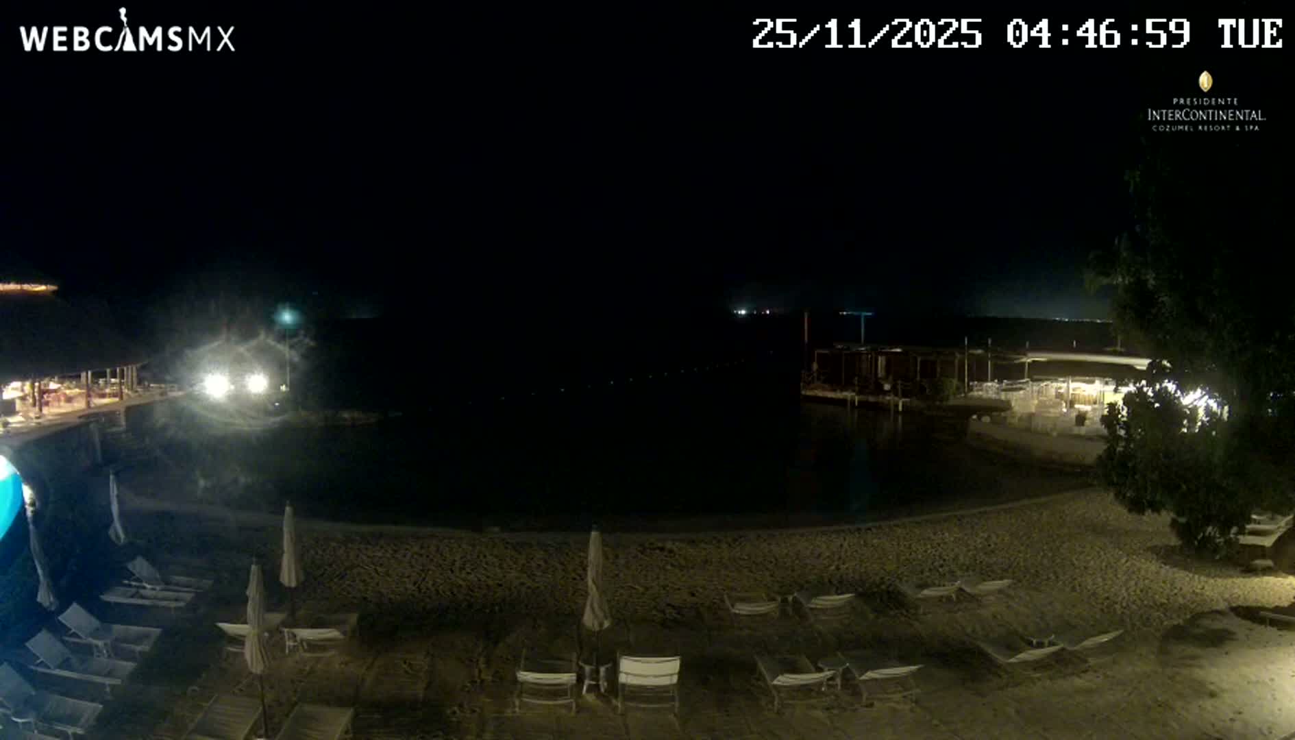 The image captures a dark night scene of a resort beach with lounge chairs and umbrellas in the foreground, illuminated buildings and structures along a waterfront, and calm, dark water under a clear sky.