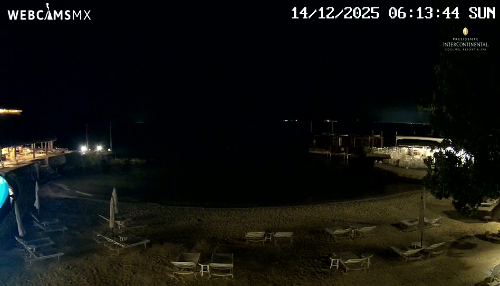The image captures a dark night scene of a resort beach with lounge chairs and umbrellas in the foreground, illuminated buildings and structures along a waterfront, and calm, dark water under a clear sky.