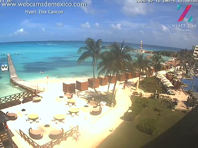 A beachfront resort with a patio seating area overlooks a calm, turquoise ocean under a partly cloudy sky.