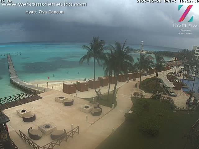 An overcast sky hangs over a beach resort with a pier extending into turquoise water, numerous palm trees lining a paved walkway leading to a sandy beach with several fire pits and lounge chairs.