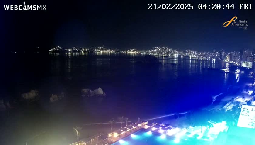 A nighttime view of a city skyline across a dark-blue ocean, with a brightly lit pool and beach area in the foreground.