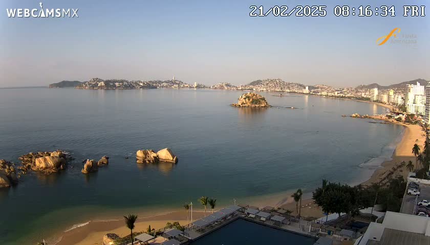 A clear, sunny day reveals a coastal city scene with a calm ocean, sandy beach, rocky outcroppings, and a distant cityscape.