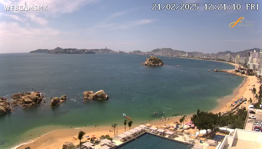 A sandy beach with clear, teal water curves around a rocky point, with a city visible in the distance under a partly sunny sky.