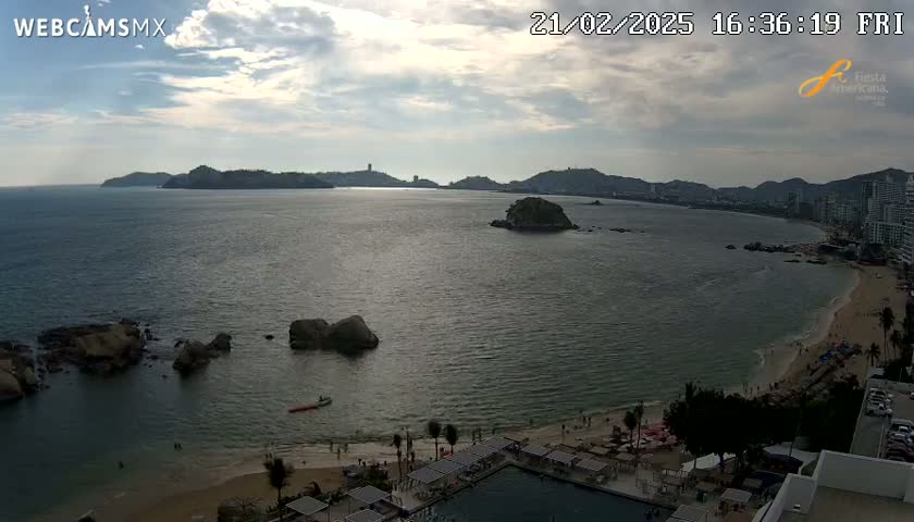 An aerial view shows a partly cloudy sky over a calm bay with a sandy beach, rocky outcroppings, and distant hills.