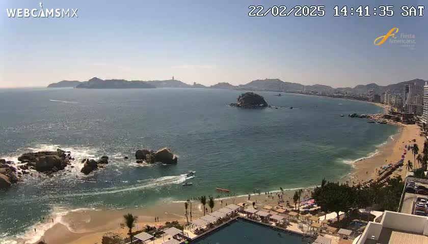 A sunny day reveals a wide, sandy beach curving along a calm bay, with rocky outcrops in the foreground and distant hills under a clear sky.