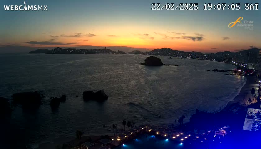 An ocean sunset view shows a city skyline with lights at dusk over dark water with rocks and a beach.