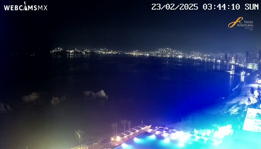 A nighttime view of a city skyline across a dark body of water, with a brightly lit pool and beach area in the foreground.