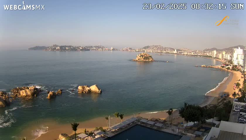 A sunny morning view of a coastal city, featuring a calm bay with rocky outcrops, a sandy beach, and a resort with a swimming pool.