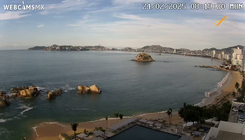 A mostly sunny day shows a coastal scene with calm ocean water, a sandy beach, rocky outcrops near the shore, a small island in the bay, and a distant city skyline.