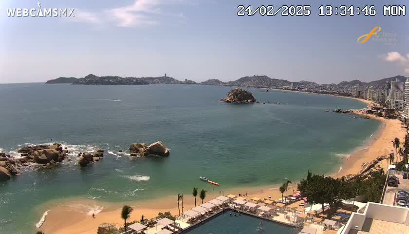 A sunny coastal scene shows a long sandy beach curving around a tranquil bay, with rocky outcrops in the foreground and a distant city skyline visible across the water.