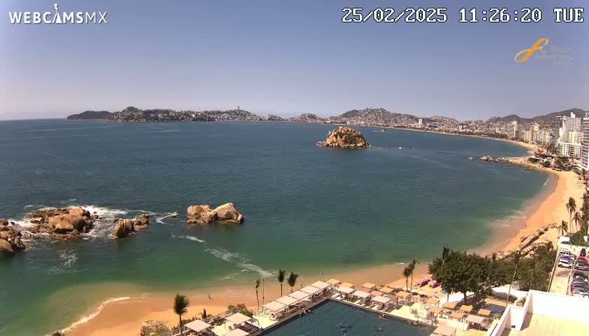A sunny day reveals a coastal city with a sandy beach, calm ocean water, and rocky outcrops, all overlooked by a resort with a pool.