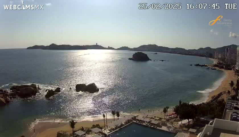 A sunny, aerial view shows a calm ocean bay with a sandy beach, rocky outcroppings, and buildings along the coastline.