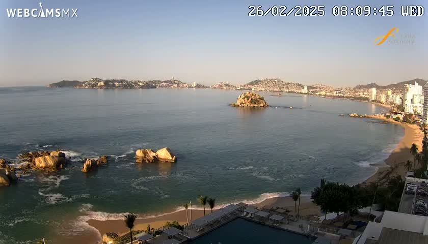 A coastal city is seen across a calm bay under a clear, bright sky, with a sandy beach and several rock formations visible in the foreground.