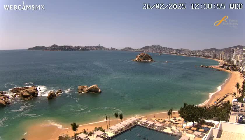 An aerial view shows a sandy beach curving around a turquoise ocean, with rocky outcrops in the water and a city visible in the distance under a clear, sunny sky.