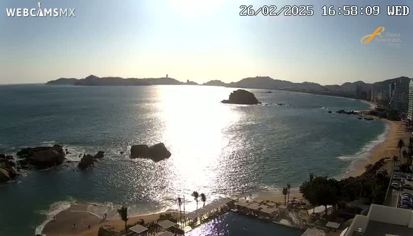 A sunny, daytime view of a beach with calm ocean water, rocky outcrops, and a resort with a pool in the foreground, backed by hills and buildings in the distance.