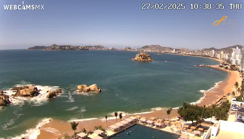 A sunny coastal scene shows a sandy beach with calm waves lapping the shore, rocky outcrops in the water, a resort with a pool, and a city in the distance.