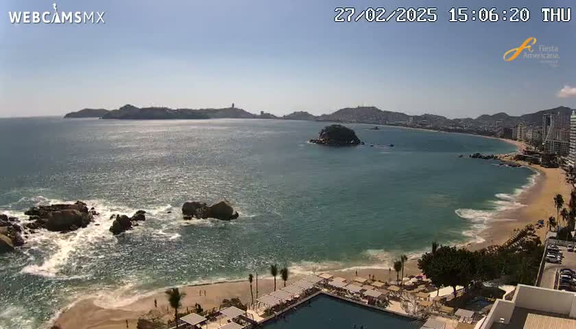 A sunny day at a beach with turquoise water, waves crashing on rocks and shore, and buildings along the coast in the distance.