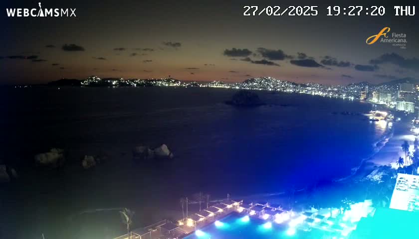 A nighttime view of a city skyline across a bay, with a beach and illuminated pool in the foreground under a partly cloudy sky.