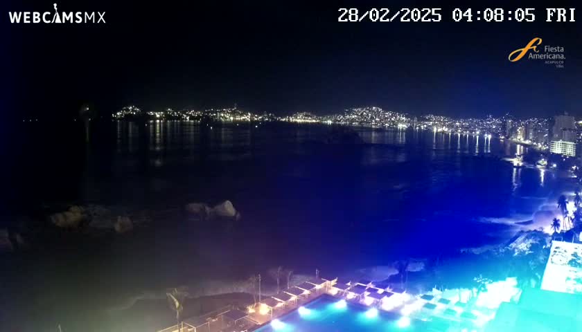 A nighttime view of a city skyline across a dark body of water, with a brightly lit pool and beach area in the foreground.