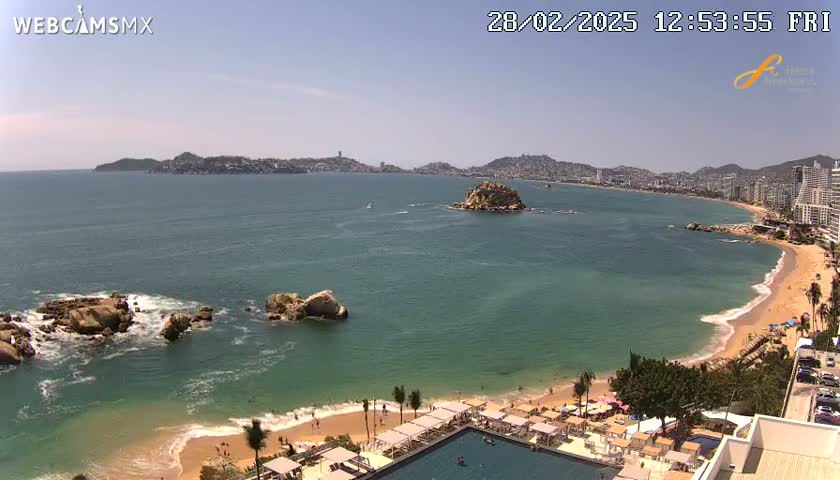 A sunny aerial view shows a calm, teal ocean lapping a sandy beach with beach umbrellas and lounge chairs, with a city and distant hills visible in the background.