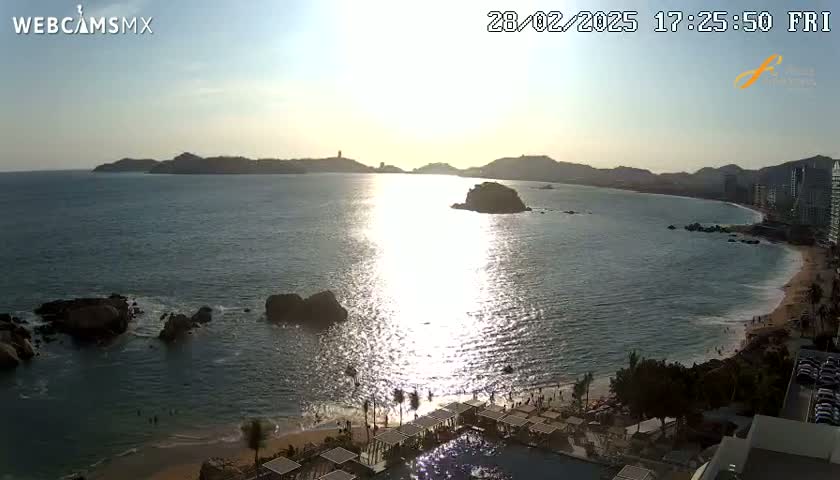 A sunny, late afternoon view of a calm ocean bay with a beach, rocky outcrops, and distant hills.