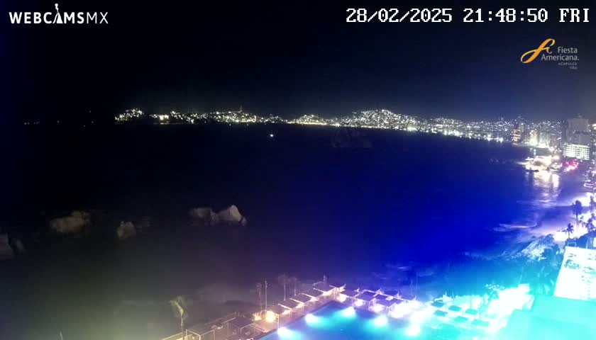 A nighttime coastal scene shows a city skyline across a dark body of water, illuminated by lights, with a brightly lit pool and surrounding structures in the foreground.