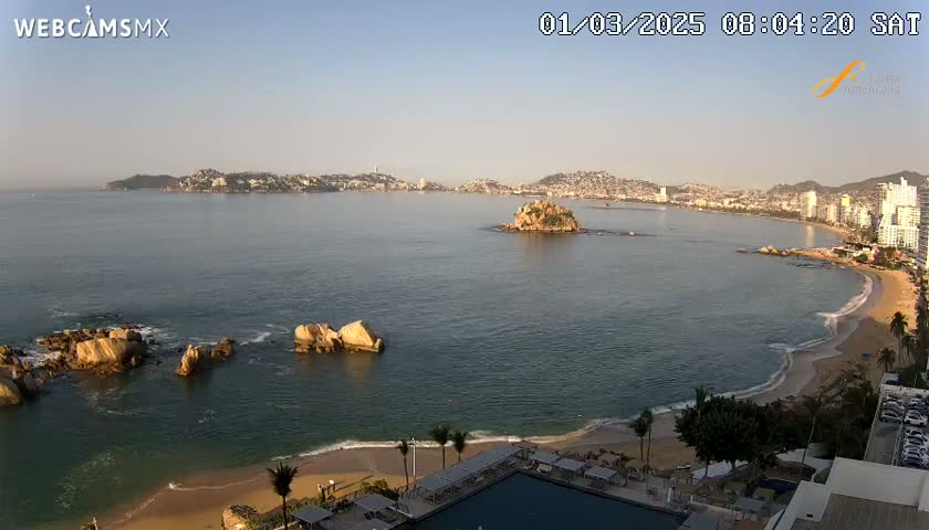 A calm, sunny ocean bay with a sandy beach, rocky outcrops, and a distant city skyline is seen from an elevated vantage point.