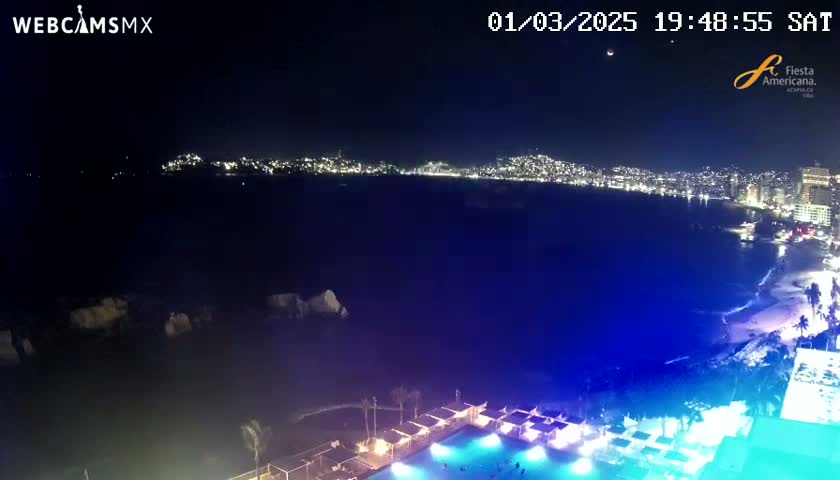 A nighttime view of a city's illuminated coastline across a dark body of water, with a pool area and beach visible in the foreground.