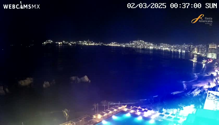 A nighttime, coastal view shows a city skyline across a dark body of water, with a brightly lit pool and cabanas in the foreground.