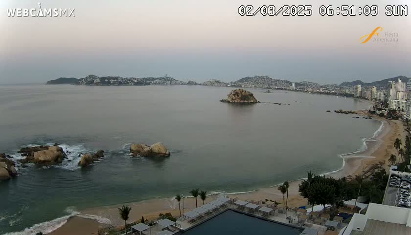 An expansive coastal view at dawn shows a calm bay with a small island, a sandy beach with gentle waves, and a resort with a pool in the foreground.