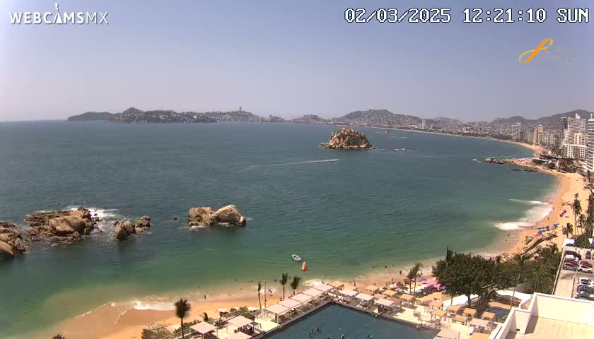 A sunny day reveals a coastal scene with a sandy beach, calm ocean waters, rocky outcrops, and a distant cityscape.
