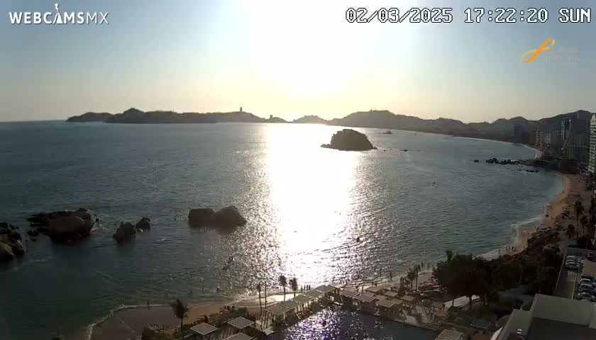 A sunny, late afternoon view of a calm bay with rocky islands, a sandy beach, and resort buildings.