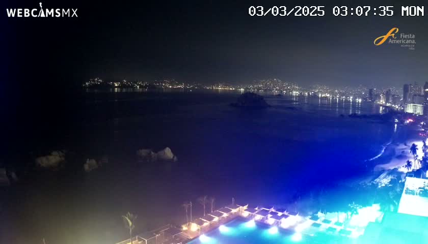 A nighttime view of a city skyline across a dark body of water, with a brightly lit pool and surrounding structures in the foreground.