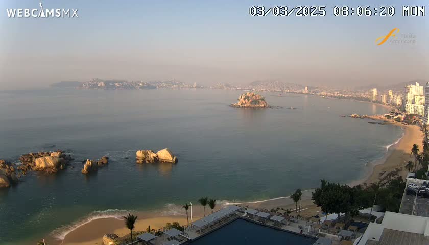 A hazy sunrise illuminates a coastal city with a sandy beach and calm ocean, featuring several rocky outcrops and a small island in the bay.